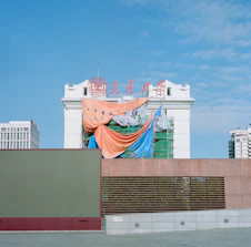 A partially constructed or renovated building draped with colorful tarps, including orange, blue, and striped sheets. The building features traditional architectural elements and has red signage on top against a clear blue sky. Modern urban structures are visible in the background.