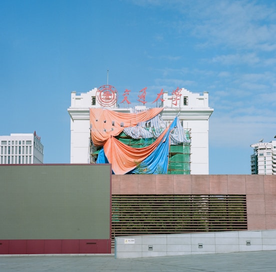 A partially constructed or renovated building draped with colorful tarps, including orange, blue, and striped sheets. The building features traditional architectural elements and has red signage on top against a clear blue sky. Modern urban structures are visible in the background.