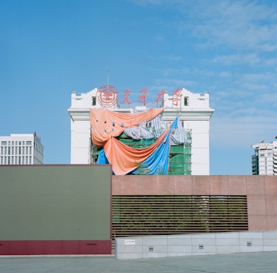 A partially constructed or renovated building draped with colorful tarps, including orange, blue, and striped sheets. The building features traditional architectural elements and has red signage on top against a clear blue sky. Modern urban structures are visible in the background.