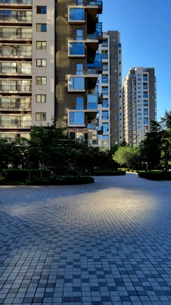 High-rise residential buildings with modern architectural features stand against a clear blue sky. The facades include balconies and large windows reflecting sunlight. In the foreground, a patterned stone plaza is shaded by surrounding trees and shrubs, creating contrast between light and shadow.
