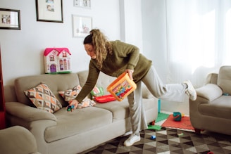 A person is tidying up a living room, holding a container of colorful toy blocks while picking up a toy car from a couch. The room has beige couches, patterned cushions, framed photos on the wall, and a toy dollhouse in the corner. The floor is covered with a geometric-patterned rug.