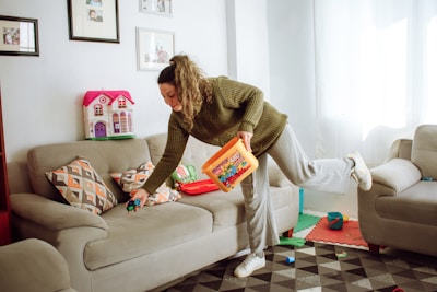 A person is tidying up a living room, holding a container of colorful toy blocks while picking up a toy car from a couch. The room has beige couches, patterned cushions, framed photos on the wall, and a toy dollhouse in the corner. The floor is covered with a geometric-patterned rug.
