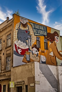 A colorful mural painted on the side of a building depicts a whimsical scene with cartoon-like characters involved in a baking theme. The artwork features a tall figure pouring flour, a child mixing ingredients in a bowl, and various kitchen items such as a mixing bowl and utensils. The mural includes a speech bubble with Hungarian text.