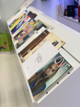 A variety of magazines are displayed on a slanted, white magazine rack. The magazines feature colorful covers with different themes and titles. A prominent image on one of the covers shows a close-up of a woman's face.