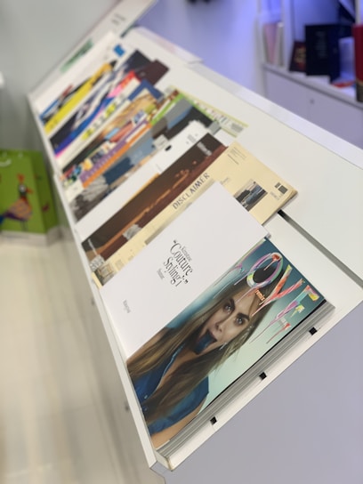A variety of magazines are displayed on a slanted, white magazine rack. The magazines feature colorful covers with different themes and titles. A prominent image on one of the covers shows a close-up of a woman's face.