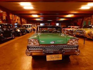 A collection of vintage cars is displayed in a dimly lit showroom. In the foreground, a green Ford Fairlane from 1959 is prominently parked, its classic chrome details gleaming under the artificial lights. Surrounding it are various other antique automobiles lined up along the walls, exhibiting a range of colors and styles.