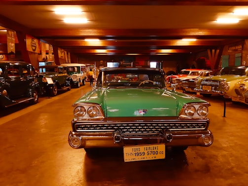 A collection of vintage cars is displayed in a dimly lit showroom. In the foreground, a green Ford Fairlane from 1959 is prominently parked, its classic chrome details gleaming under the artificial lights. Surrounding it are various other antique automobiles lined up along the walls, exhibiting a range of colors and styles.
