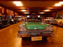 A collection of vintage cars is displayed in a dimly lit showroom. In the foreground, a green Ford Fairlane from 1959 is prominently parked, its classic chrome details gleaming under the artificial lights. Surrounding it are various other antique automobiles lined up along the walls, exhibiting a range of colors and styles.