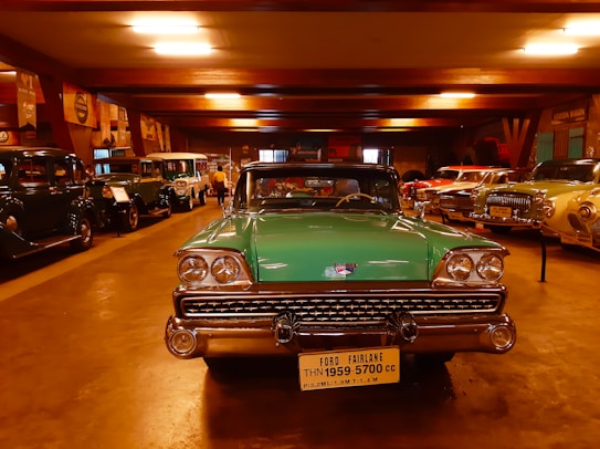 A collection of vintage cars is displayed in a dimly lit showroom. In the foreground, a green Ford Fairlane from 1959 is prominently parked, its classic chrome details gleaming under the artificial lights. Surrounding it are various other antique automobiles lined up along the walls, exhibiting a range of colors and styles.
