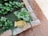 A garden scene with lush green leaves growing in a small patch bordered by bricks. A pair of yellow gardening gloves is placed on the brick edging. The ground around the garden bed is covered with reddish-brown gravel.