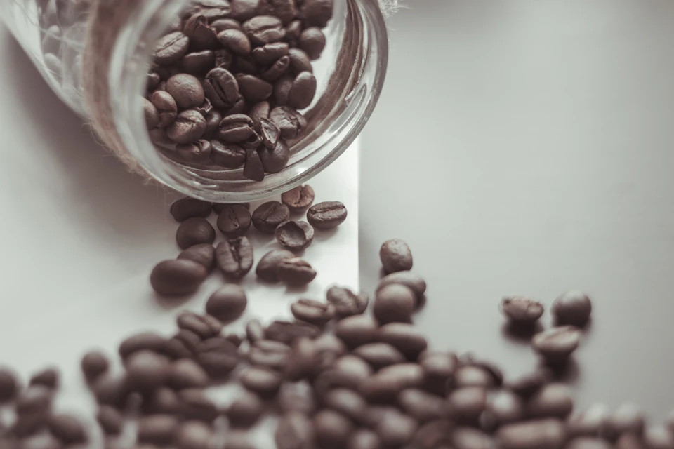 Coffee beans spill from a jar onto a white surface, creating a brown ground pattern