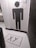 The image depicts a bathroom entrance with a tile design featuring a large, stylized person icon made from black hexagonal tiles against a white hexagonal tile background. The floor also consists of white hexagonal tiles with a black 'XX' symbol inside a rectangular border near the entrance. A trash can is visible on the left side.