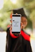 A person wearing a graduation cap and gown holds a smartphone displaying the text 'classe of 2022' with a graduation cap graphic. The background is blurred, emphasizing the phone and the message.