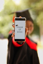 A person wearing a graduation cap and gown holds a smartphone displaying the text 'classe of 2022' with a graduation cap graphic. The background is blurred, emphasizing the phone and the message.