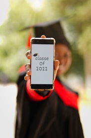 A person wearing a graduation cap and gown holds a smartphone displaying the text 'classe of 2022' with a graduation cap graphic. The background is blurred, emphasizing the phone and the message.