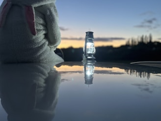 A plush toy wearing a hood sits atop a reflective surface near a lantern with its light on. The sky in the background is dusk, showing a gradient of light blue to deep orange, with silhouetted trees on the horizon.