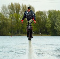 A person wearing a black jacket and a hat is hydrofoiling above a body of water. Surrounding the scene are tall trees with green and brown leaves, indicating a natural setting. The person appears to be hovering above the water with a concentrated expression.