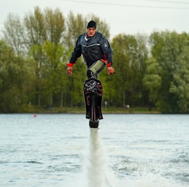 A person wearing a black jacket and a hat is hydrofoiling above a body of water. Surrounding the scene are tall trees with green and brown leaves, indicating a natural setting. The person appears to be hovering above the water with a concentrated expression.