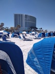 A beach scene with numerous blue beach cabanas and lounge chairs spread across the white sandy beach. A modern, curved high-rise building stands in the background, flanked by palm trees. The sky is clear and blue, creating a serene and inviting atmosphere.