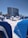A beach scene with numerous blue beach cabanas and lounge chairs spread across the white sandy beach. A modern, curved high-rise building stands in the background, flanked by palm trees. The sky is clear and blue, creating a serene and inviting atmosphere.