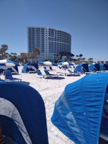 A beach scene with numerous blue beach cabanas and lounge chairs spread across the white sandy beach. A modern, curved high-rise building stands in the background, flanked by palm trees. The sky is clear and blue, creating a serene and inviting atmosphere.