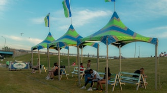A group of colorful, triangular canopy tents with flags on top are set up on a grassy field. In the foreground, several benches are placed under the tents, with people sitting and relaxing on them. A few individuals are walking or sitting on the grass in the background. The sky is clear with some clouds, and there is a bridge and a road with light poles in the background.