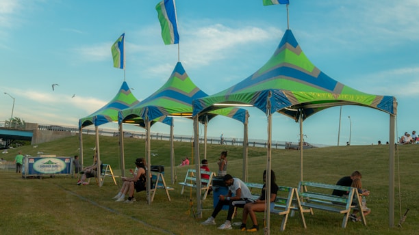 A group of colorful, triangular canopy tents with flags on top are set up on a grassy field. In the foreground, several benches are placed under the tents, with people sitting and relaxing on them. A few individuals are walking or sitting on the grass in the background. The sky is clear with some clouds, and there is a bridge and a road with light poles in the background.
