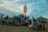 A bustling fairground scene with people gathered around a colorful booth filled with stuffed animals and prizes. In the background, a tall illuminated structure and a carousel are visible. Individuals are interacting, walking, and sitting on benches, creating a lively and vibrant atmosphere.