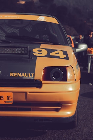 A close-up view of the front portion of an orange Renault car displaying the number 94. The car is parked outdoors, with some people visible in the background. The lighting suggests late afternoon or early evening, creating a subtle contrast between the car and the darker surroundings.