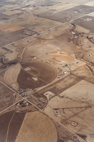 An aerial view of expansive farmland divided into various rectangular and circular plots. The fields vary in color and size, indicating different stages of cultivation or use. Sparse buildings and farmhouses are scattered across the landscape, connected by a network of dirt roads. The land appears dry and barren, possibly indicating an arid climate.