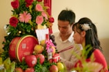 A couple dressed formally engages in an intimate moment, surrounded by an arrangement of vibrant flowers, fruits, and a decorative red box. The setting suggests a ceremonial atmosphere, with symbolic elements like incense sticks in the man's hand.