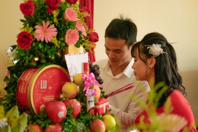 A couple dressed formally engages in an intimate moment, surrounded by an arrangement of vibrant flowers, fruits, and a decorative red box. The setting suggests a ceremonial atmosphere, with symbolic elements like incense sticks in the man's hand.