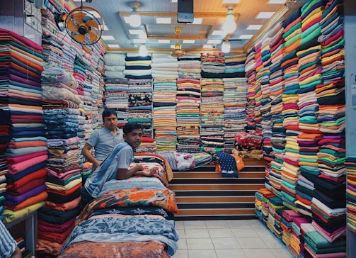 a couple of men sitting on a bed with a large pile of colorful fabric