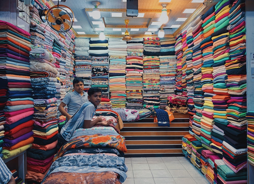 a couple of men sitting on a bed with a large pile of colorful fabric