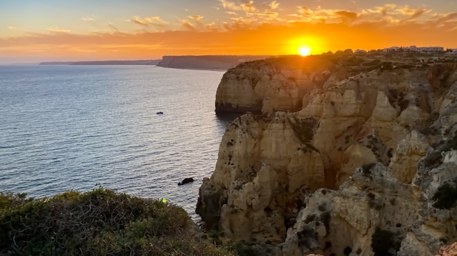 A vibrant sunset casts a warm orange glow over the cliff-lined coast. The sky features scattered clouds illuminated by the sun, creating a dramatic effect. The ocean extends calmly into the horizon, with a small boat visible on the water. The rugged cliffs display intricate geological formations, adding depth and interest to the scene.