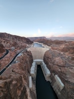 A large concrete dam is situated between rocky canyons with a narrow body of water at its base. The structure is imposing, with roads and infrastructure visible on either side, suggesting an industrial setting. The landscape around it is arid and mountainous, with a clear blue sky above.