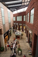 A modern indoor shopping center with large windows, brick and stone walls, and multiple stores lining the sides. There is a small kiosk in the center named 'Caba Mitt' and various small tables set up with products. Several people are walking around, browsing or shopping.