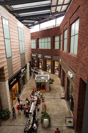 A modern indoor shopping center with large windows, brick and stone walls, and multiple stores lining the sides. There is a small kiosk in the center named 'Caba Mitt' and various small tables set up with products. Several people are walking around, browsing or shopping.