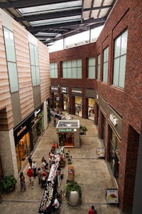A modern indoor shopping center with large windows, brick and stone walls, and multiple stores lining the sides. There is a small kiosk in the center named 'Caba Mitt' and various small tables set up with products. Several people are walking around, browsing or shopping.
