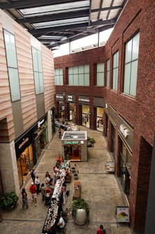 A modern indoor shopping center with large windows, brick and stone walls, and multiple stores lining the sides. There is a small kiosk in the center named 'Caba Mitt' and various small tables set up with products. Several people are walking around, browsing or shopping.