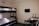 A small room contains a bunk bed with white bedding against a beige wall. Adjacent to the bed is a table holding a kettle, cups, a bowl, and two large water bottles. A flat-screen TV is mounted on the wall above. A jacket is draped over a chair nearby.