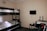 A small room contains a bunk bed with white bedding against a beige wall. Adjacent to the bed is a table holding a kettle, cups, a bowl, and two large water bottles. A flat-screen TV is mounted on the wall above. A jacket is draped over a chair nearby.