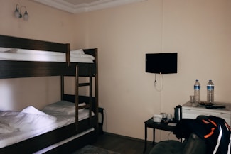 A small room contains a bunk bed with white bedding against a beige wall. Adjacent to the bed is a table holding a kettle, cups, a bowl, and two large water bottles. A flat-screen TV is mounted on the wall above. A jacket is draped over a chair nearby.