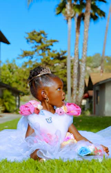 a little girl in a white dress sitting on grass with trees and a building in the background