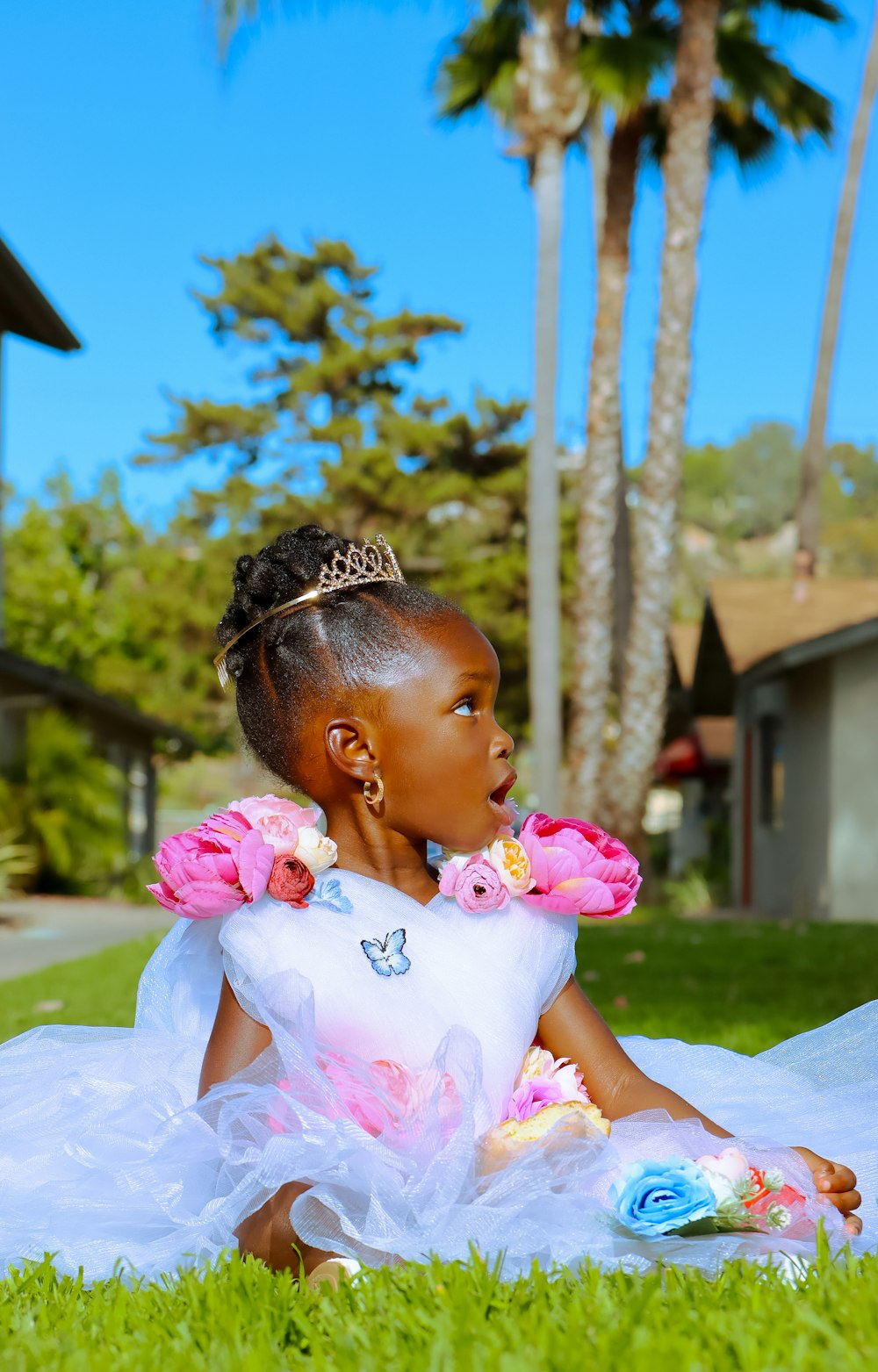 a little girl in a white dress sitting on grass with trees and a building in the background