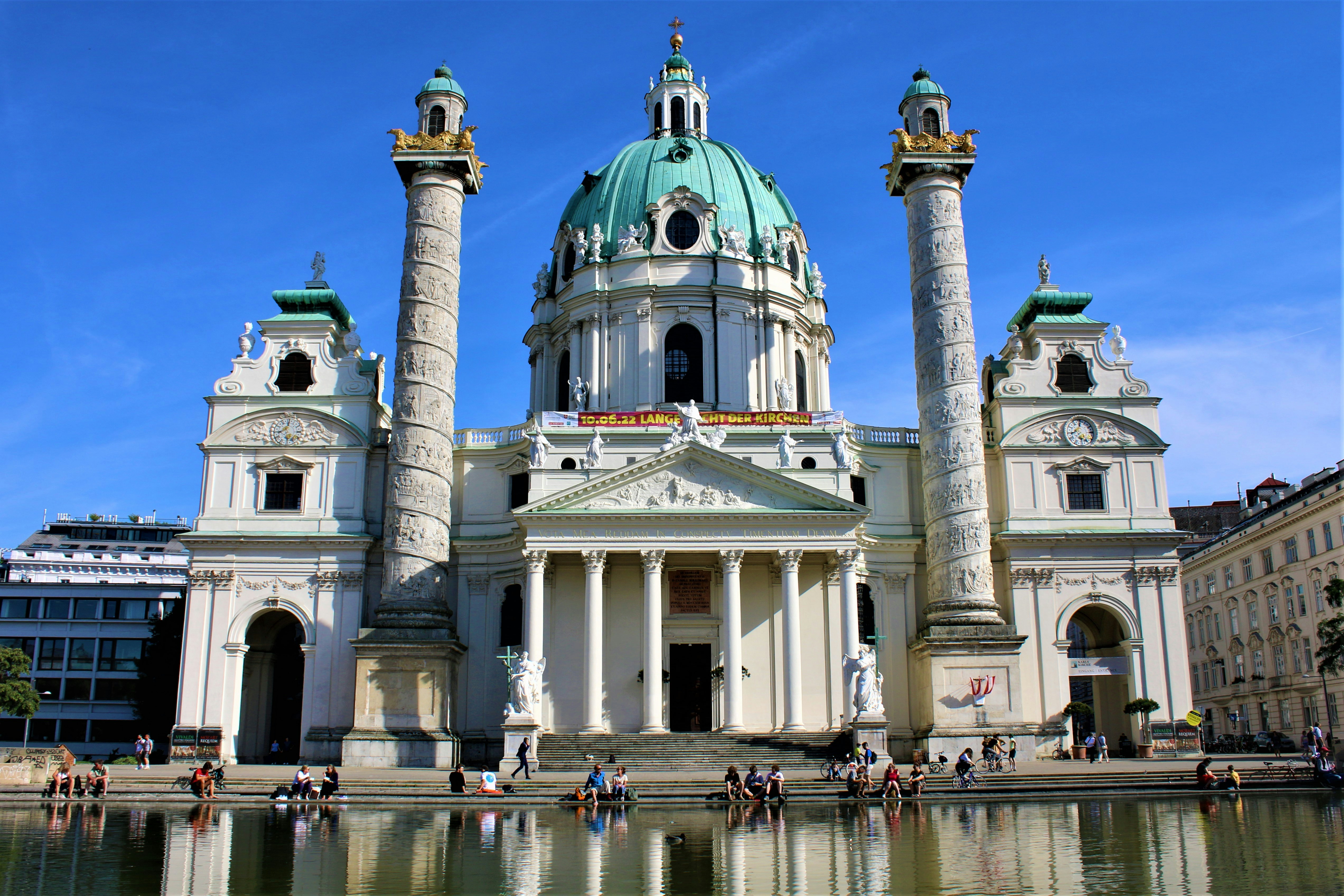 un grand bâtiment blanc avec un toit en forme de dôme et des piliers près d’un plan d’eau avec Karlskirche en arrière-plan