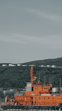 A bright orange industrial ship is docked near a lush green hillside under a clear sky. The ship, named Stakhanovets, features multiple antennas and equipment on its deck. In the distance, several white storage tanks are visible atop the hill.