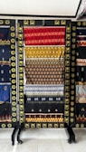 A collection of vibrant woven textiles is displayed on a black rack, each featuring intricate geometric patterns in red, yellow, and various colors. The textiles appear handcrafted with traditional designs, surrounded by a decorative frame with gold accents.