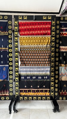 A collection of vibrant woven textiles is displayed on a black rack, each featuring intricate geometric patterns in red, yellow, and various colors. The textiles appear handcrafted with traditional designs, surrounded by a decorative frame with gold accents.
