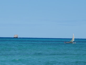 A small sailboat with a white sail travels across the turquoise ocean. In the background, a rocky islet is visible under a clear blue sky.